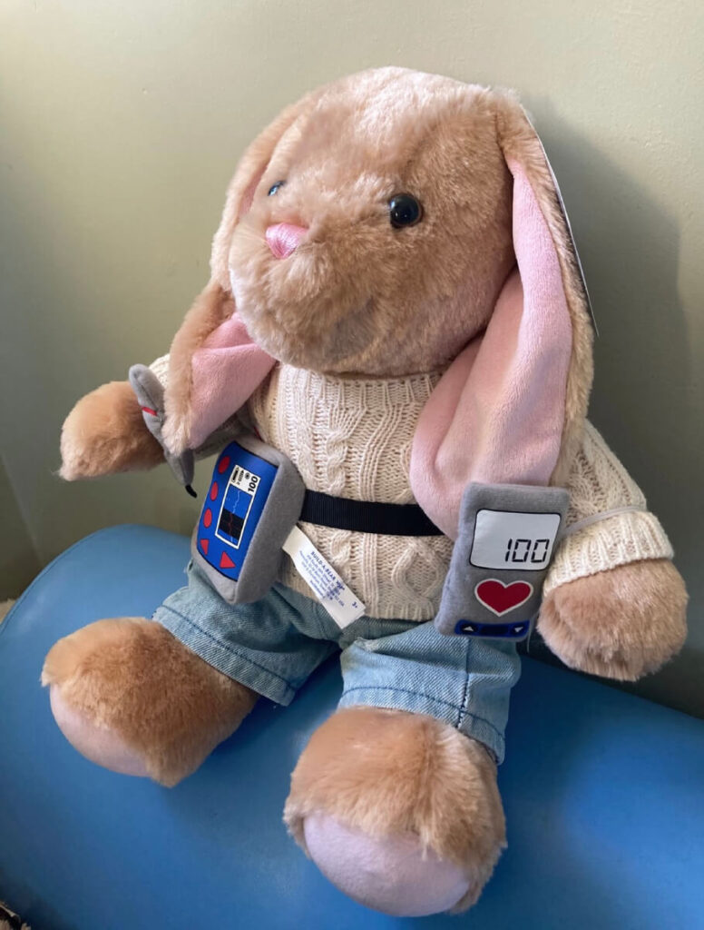 The image shows a plush toy in the form of a light brown bunny with floppy pink ears. The bunny is dressed in a white knitted sweater and blue denim pants. It has a small black belt around its waist, attached to which are two gray, rectangular devices. One of the devices resembles a monitor with a digital display reading "100" and a red heart icon below it. The other device has a blue screen and red buttons, giving the impression of a handheld gaming or electronic device. The bunny is seated on a blue cushion with a light green wall in the background.